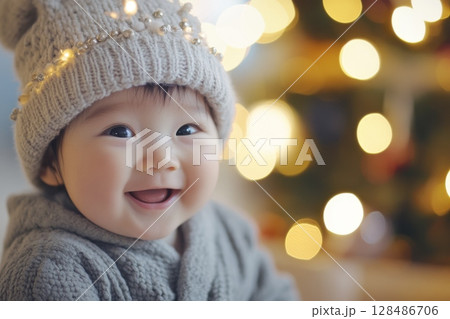 Joyful Asian Baby in Wool Hat Surrounded by Twinkling Christmas Lights and Decor. Happy cheerful toddler child laughing on colorful festive Christmas background among Xmas Tree. Copy Space 128486706