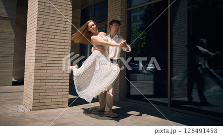 Joyful woman dancing in arms of partner on urban street with sunlit reflection in glass wall Joyful woman dancing in arms of partner on urban street with sunlit reflection in glass wall 128491458