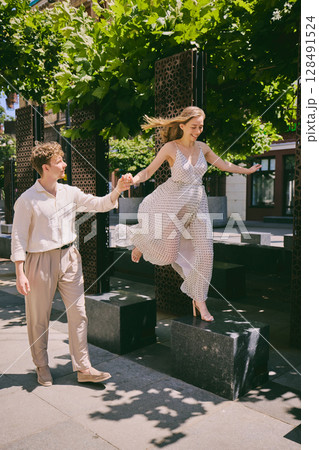Cheerful young woman jumping playfully while holding man hand in sunny modern city setting 128491524