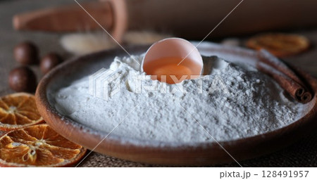 Food still life of organic food ingredients for homemade baking with white flour and egg yolk inside half of broken egg shell. Flour pile in traditional wooden bowl. Dried orange slices and cinnamon 128491957