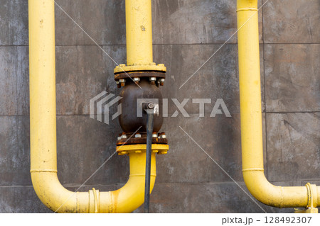 Yellow industrial pipes connect to a valve on a textured concrete wall, highlighting infrastructure design and maintenance work during the day 128492307