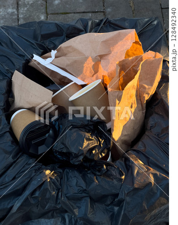 Coffee cups and crumpled paper waste overflow from a black trash bag, illuminated by warm late afternoon sunlight in a city environment 128492340