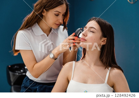 Beauty salon worker is examining skin of the woman by using a device with light and lens Beauty salon worker is examining skin of the woman by using a device with light and lens 128492426
