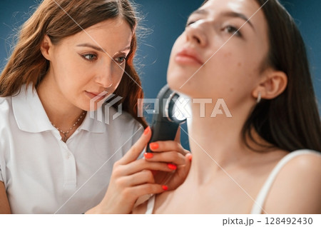 Beauty salon worker is examining skin of the woman by using a device with light and lens 128492430