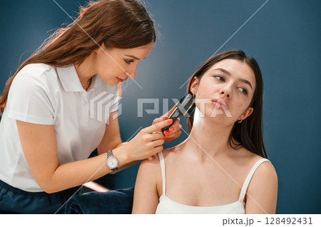Against blue wall. Beauty salon worker is examining skin of the woman by using a device with light and lens 128492431