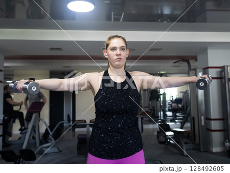 Ordinary woman in the gym doing exercises with dumbbells. Workout for weight loss. Body strengthening and healthy lifestyle Ordinary woman in the gym doing exercises with dumbbells. Workout for weight loss. Body strengthening and healthy lifestyle 128492605