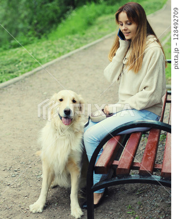 Young woman owner talking on phone with Golden Retriever dog sitting on bench in summer park 128492709