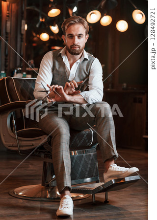 Holding scissors. Handsome barber is in the salon Holding scissors. Handsome barber is in the salon 128492721