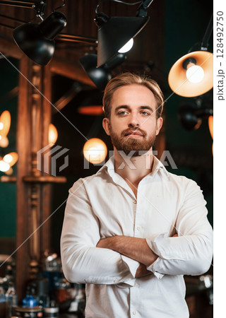 Standing with arms crossed. Handsome barber is in the salon Standing with arms crossed. Handsome barber is in the salon 128492750