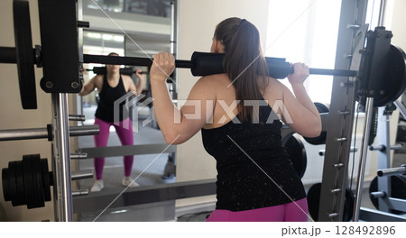Plus size woman doing exercises on Smith Machine. Squats on a simulator with a barbell. Workout for weight loss Plus size woman doing exercises on Smith Machine. Squats on a simulator with a barbell. Workout for weight loss 128492896