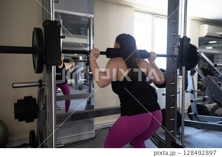 Plus size woman doing exercises on Smith Machine. Squats on a simulator with a barbell. Workout for weight loss Plus size woman doing exercises on Smith Machine. Squats on a simulator with a barbell. Workout for weight loss 128492897