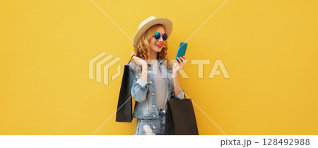 Beautiful happy young woman holding phone with black shopping bag, summer hat on yellow background 128492988