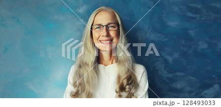 Portrait of happy smiling confident mature woman with eyeglasses looks at camera on blue background 128493033