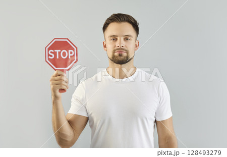 Young confident man holding red stop sign in hands standing indoors on a gray studio background Young confident man holding red stop sign in hands standing indoors on a gray studio background 128493279