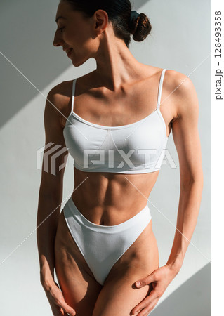 Standing in the studio with shadows. Young woman with slim body type is in fitness clothes Standing in the studio with shadows. Young woman with slim body type is in fitness clothes 128493358