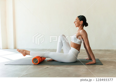 Orange colored foam roller. Young woman with slim body type is in fitness clothes in the studio 128493377