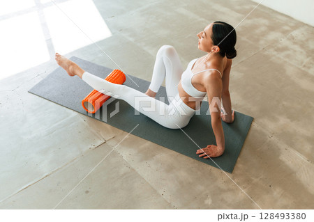 With orange colored foam roller. Young woman with slim body type is in fitness clothes in the studio With orange colored foam roller. Young woman with slim body type is in fitness clothes in the studio 128493380