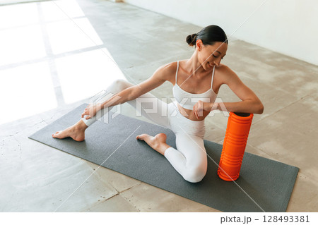 With orange colored foam roller. Young woman with slim body type is in fitness clothes in the studio 128493381