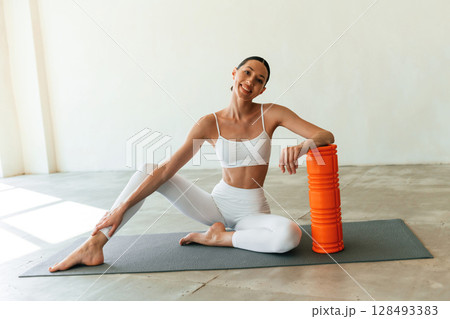 With orange colored foam roller. Young woman with slim body type is in fitness clothes in the studio With orange colored foam roller. Young woman with slim body type is in fitness clothes in the studio 128493383