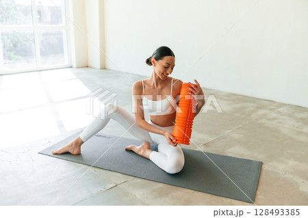 Sitting with foam roller. Young woman with slim body type is in fitness clothes in the studio 128493385