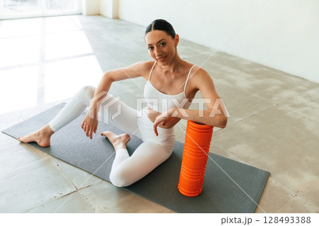 Sitting with foam roller. Young woman with slim body type is in fitness clothes in the studio Sitting with foam roller. Young woman with slim body type is in fitness clothes in the studio 128493388