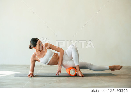 Using foam roller for exercises. Young woman with slim body type is in fitness clothes in the studio Using foam roller for exercises. Young woman with slim body type is in fitness clothes in the studio 128493393
