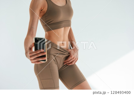 Taking a selfie, photo of abs. Young woman with slim body type is in fitness clothes in the studio 128493400