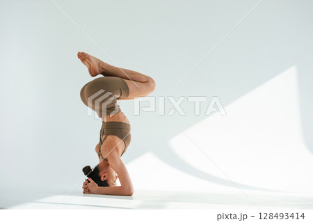 Doing head stand. Young woman with slim body type is in fitness clothes in the studio 128493414
