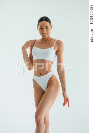 Feeling good. Standing and smiling. Young woman with slim body type is in fitness clothes in the studio Feeling good. Standing and smiling. Young woman with slim body type is in fitness clothes in the studio 128493488