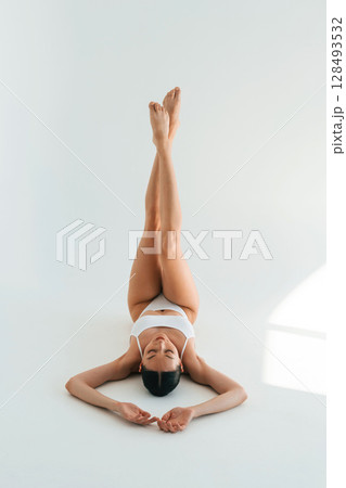 Legs are up, lying down. Young woman with slim body type is in fitness clothes in the studio Legs are up, lying down. Young woman with slim body type is in fitness clothes in the studio 128493532