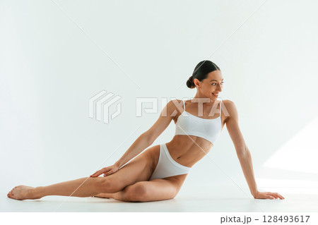 Sitting on the floor. Young woman with slim body type is in fitness clothes in the studio Sitting on the floor. Young woman with slim body type is in fitness clothes in the studio 128493617