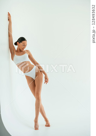 Leaning on the white surface. Young woman with slim body type is in fitness clothes in the studio Leaning on the white surface. Young woman with slim body type is in fitness clothes in the studio 128493632