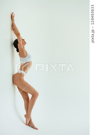 Leaning on the white surface. Young woman with slim body type is in fitness clothes in the studio Leaning on the white surface. Young woman with slim body type is in fitness clothes in the studio 128493633