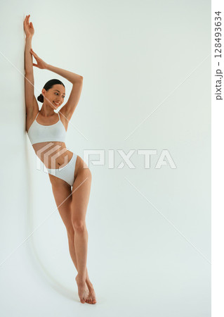 Leaning on the white surface. Young woman with slim body type is in fitness clothes in the studio 128493634