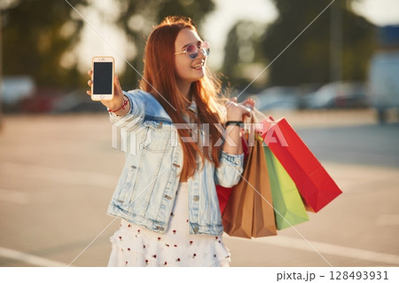 Showing smartphone that is in the hand. Beautiful woman in casual clothes is holding shopping bags, outdoors 128493931