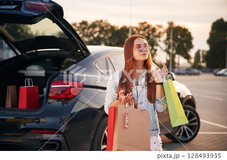 Standing near the car that is with the trunk open. Beautiful woman in casual clothes is holding shopping bags, outdoors 128493935