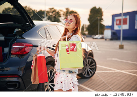 Standing near the car that is with the trunk open. Beautiful woman in casual clothes is holding shopping bags, outdoors Standing near the car that is with the trunk open. Beautiful woman in casual clothes is holding shopping bags, outdoors 128493937