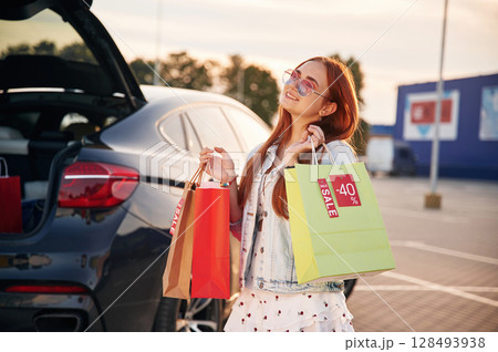Standing near the car that is with the trunk open. Beautiful woman in casual clothes is holding shopping bags, outdoors 128493938