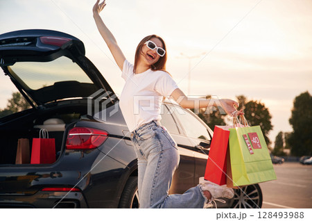 Against the car with trunk opened. Beautiful woman in casual clothes is holding shopping bags, outdoors 128493988