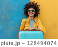 Smiling woman wearing sunglasses with blue suitcase standing against blue and yellow wall 128494074