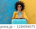 Smiling woman wearing sunglasses with blue suitcase standing against blue and yellow wall 128494075