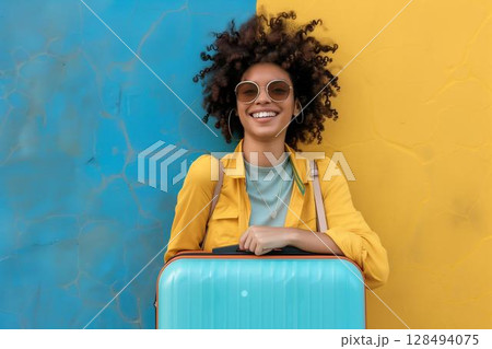 Smiling woman wearing sunglasses with blue suitcase standing against blue and yellow wall 128494075
