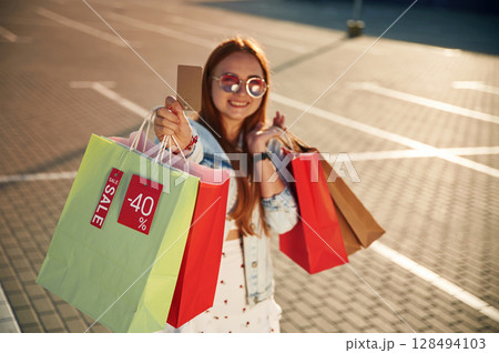 Discounts, with credit card in hands. Beautiful woman in casual clothes is holding shopping bags, outdoors 128494103
