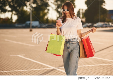 Using smartphone. Beautiful woman in casual clothes is holding shopping bags, outdoors 128494262