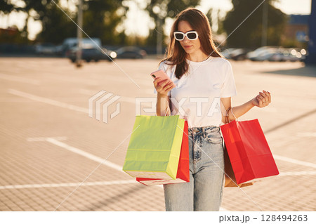 Using smartphone. Beautiful woman in casual clothes is holding shopping bags, outdoors 128494263
