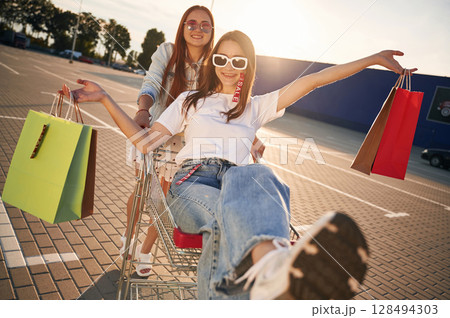 Riding in a shop cart. Two beautiful women in casual clothes are holding bags, outdoors Riding in a shop cart. Two beautiful women in casual clothes are holding bags, outdoors 128494303