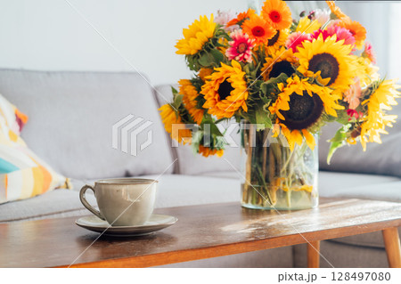 Focus on cup of hot drink and vase with huge sunflowers bouquet on wooden coffee table in modern cozy light living room with gray couch sofa. Cozy Design home decor. Summer mood, Relaxing rituals. 128497080