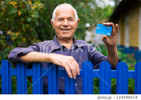 Portrait of satisfied old man 70s with gray hair smiling while holding credit card over country house background 128498414