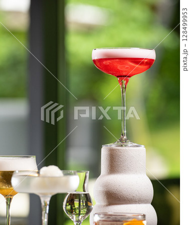 A vibrant red cocktail with a delicate white foam top is elegantly presented on a unique textured white stand, amidst other sophisticated drinks, against a lush green blurred background. 128499953