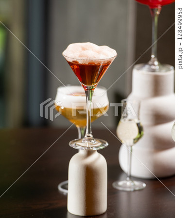 A richly colored red cocktail with a vibrant orange peel garnish is presented in a textured rocks glass, surrounded by other sophisticated drinks in a dimly lit bar setting. A richly colored red cocktail with a vibrant orange peel garnish is presented in a textured rocks glass, surrounded by other sophisticated drinks in a dimly lit bar setting. 128499958